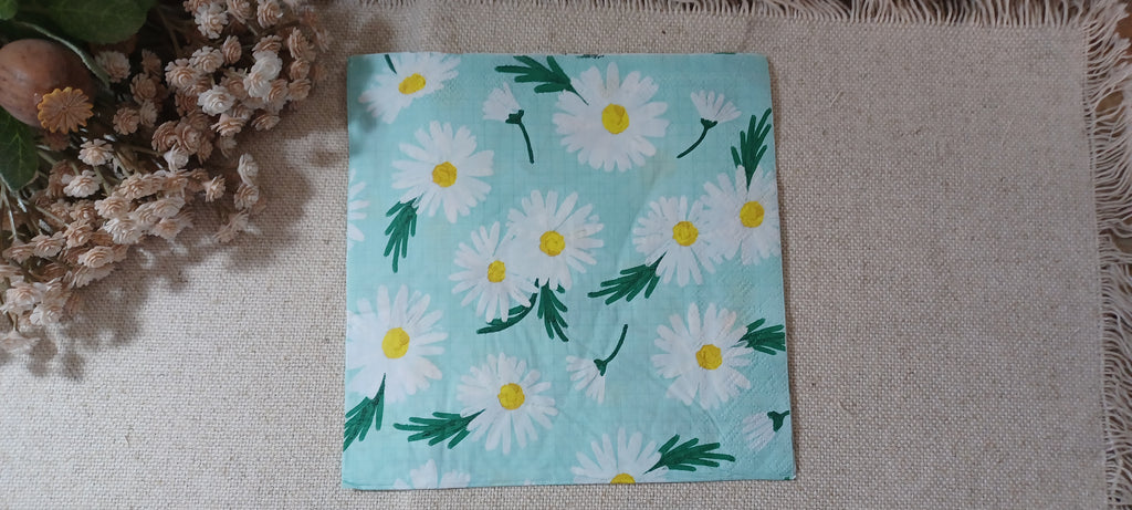 A square paper napkin with a daisy pattern on a light blue background, displayed on a beige woven mat next to dried flowers. Great for crafts