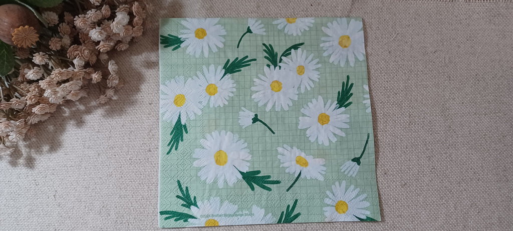 A square paper napkin featuring a daisy pattern on a light green background, laid flat on a beige textured surface next to dried flowers. Great for crafts