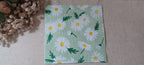A square paper napkin featuring a daisy pattern on a light green background, laid flat on a beige textured surface next to dried flowers. Great for crafts