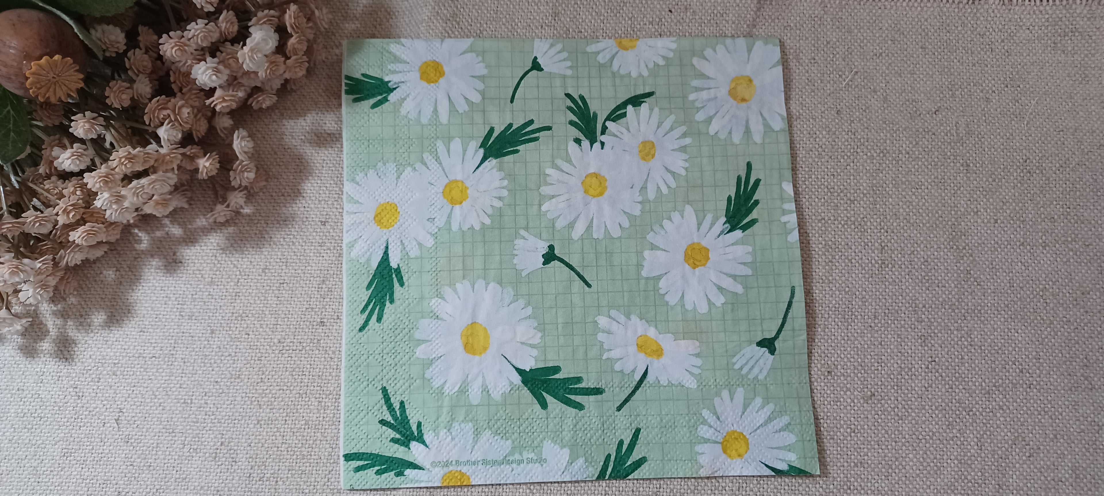 A square paper napkin featuring a daisy pattern on a light green background, laid flat on a beige textured surface next to dried flowers. Great for crafts