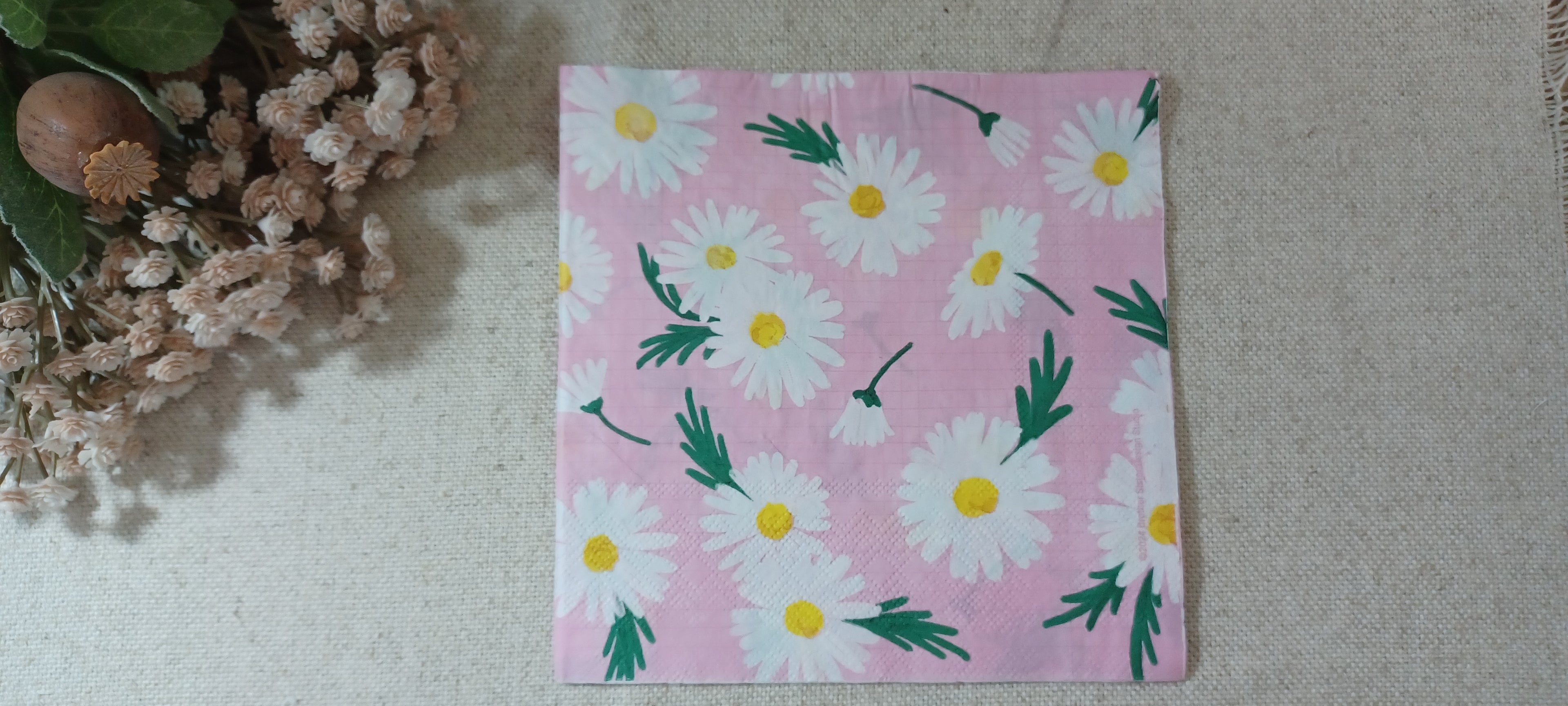 A square pink paper napkin with a white daisy floral pattern, displayed on a beige textured fabric surface next to dried flowers. Great for crafts