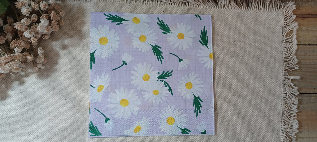 A square paper napkin featuring a daisy pattern on a light lavender background, laid flat on a beige fringed mat with dried flowers in the corner. Great for crafts