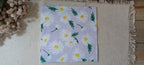 A square paper napkin featuring a daisy pattern on a light lavender background, laid flat on a beige fringed mat with dried flowers in the corner. Great for crafts