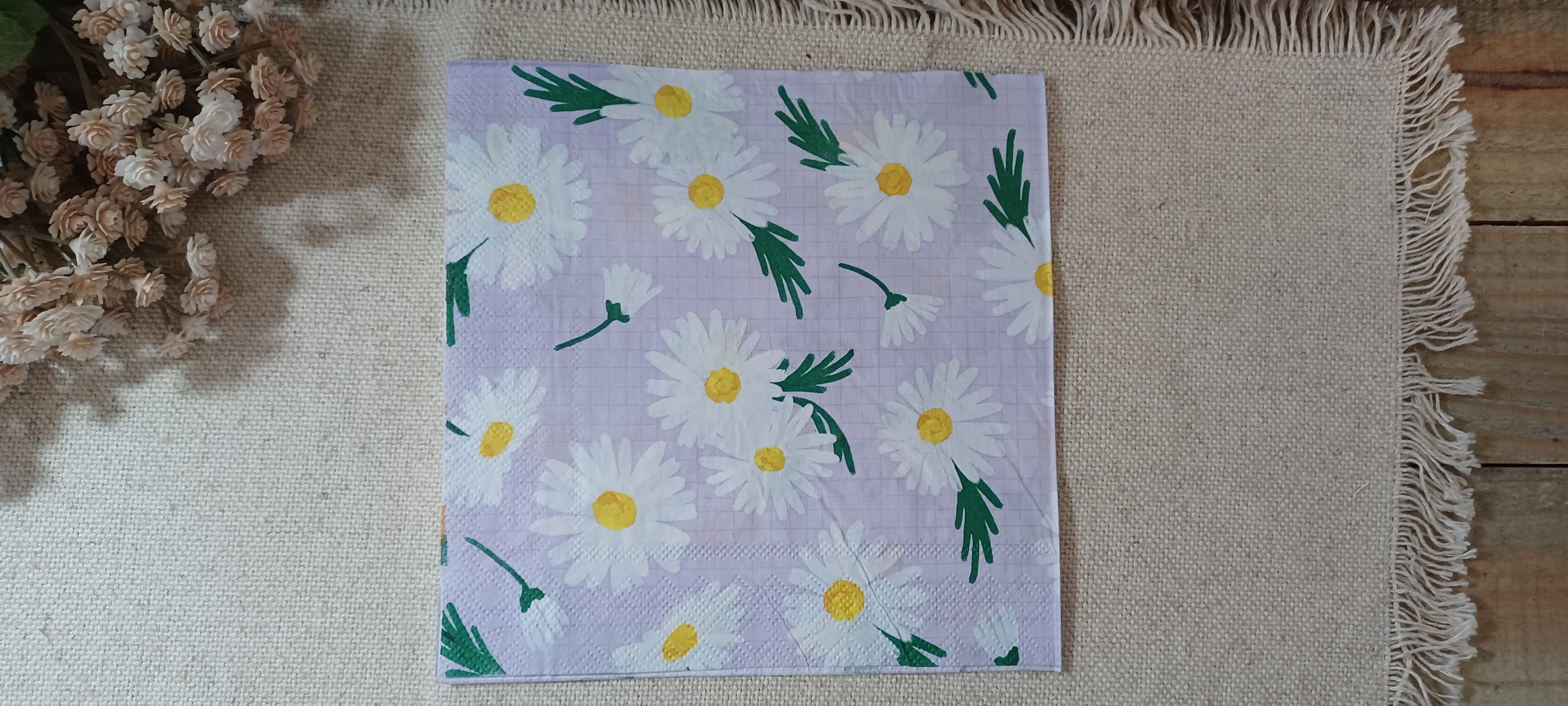 A square paper napkin featuring a daisy pattern on a light lavender background, laid flat on a beige fringed mat with dried flowers in the corner. Great for crafts
