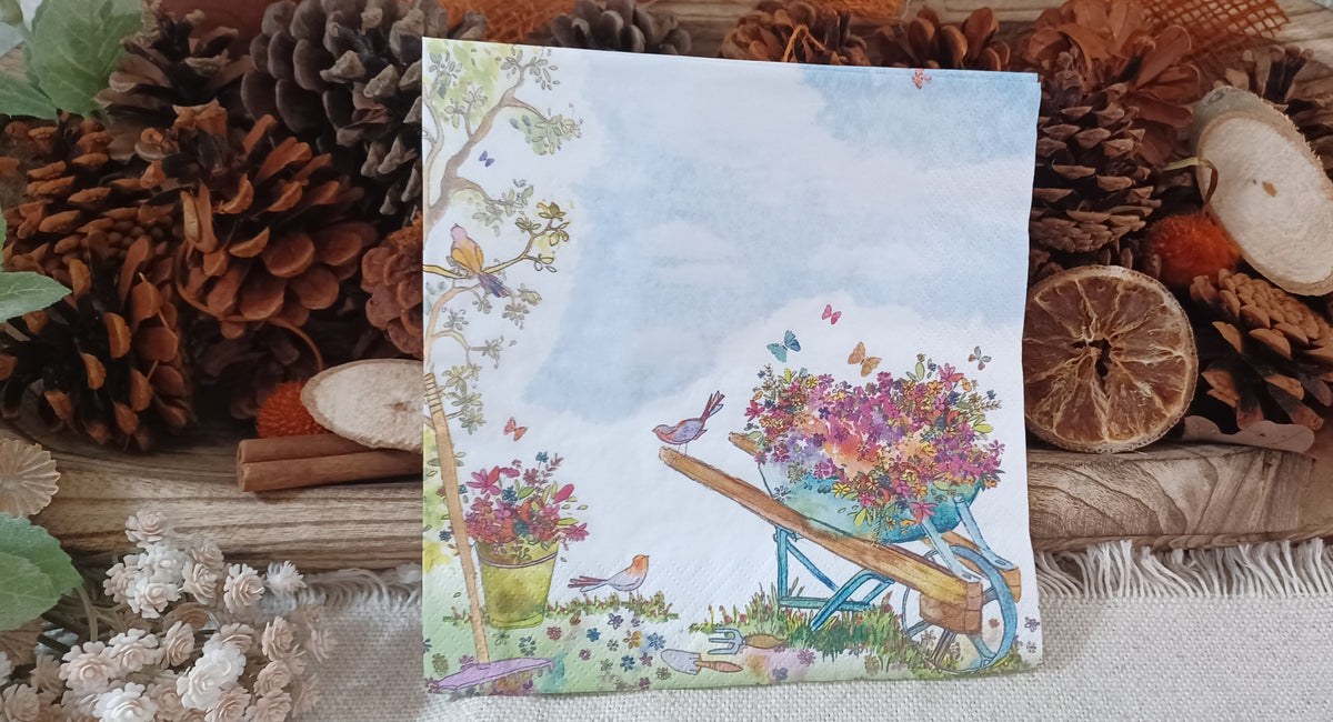 Garden-themed decorative paper napkin featuring a flower-filled wheelbarrow, birds, and butterflies displayed with rustic natural decor.