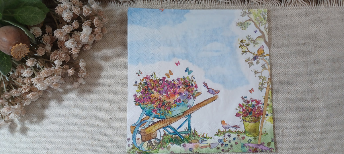 Floral wheelbarrow decoupage napkin with watercolor garden flowers, birds, butterflies, and soft blue sky in a rustic display.