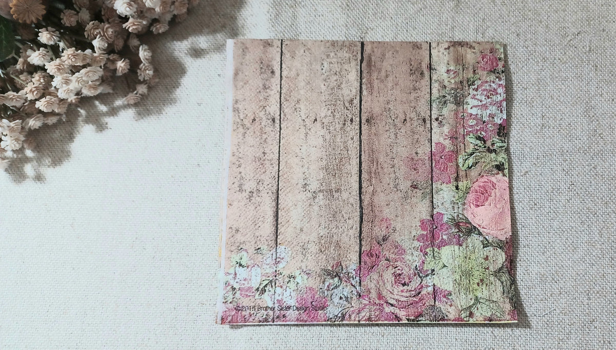 Vintage-style paper napkin with faded pink roses and greenery over a distressed brown wood plank design.