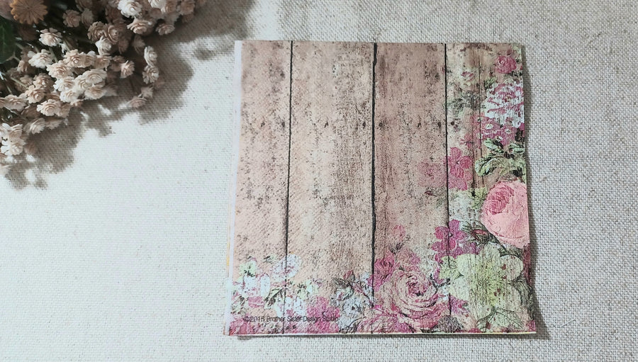 Vintage-style paper napkin with faded pink roses and greenery over a distressed brown wood plank design.