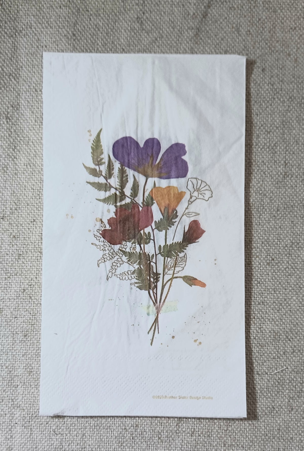 Full view of a botanical guest napkin featuring purple and rust wildflowers, golden blossoms, fern fronds and delicate line-drawn flowers.