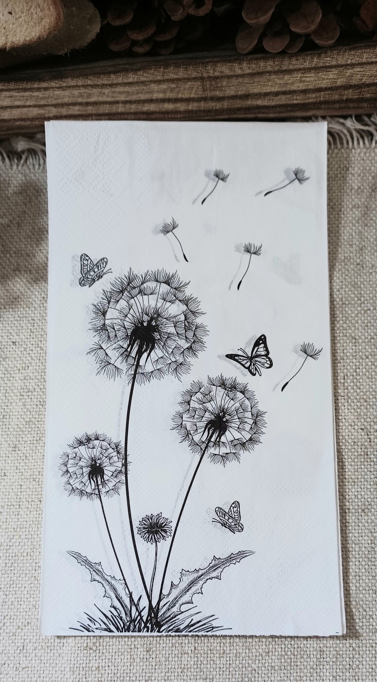 Black and white paper guest napkin featuring illustrated dandelions, floating seeds and butterflies.