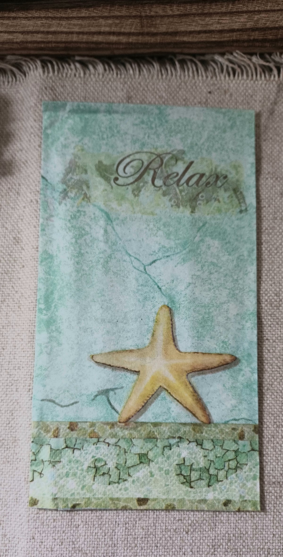 Aqua coastal paper guest napkin featuring a golden starfish, distressed background, and the word Relax in script.