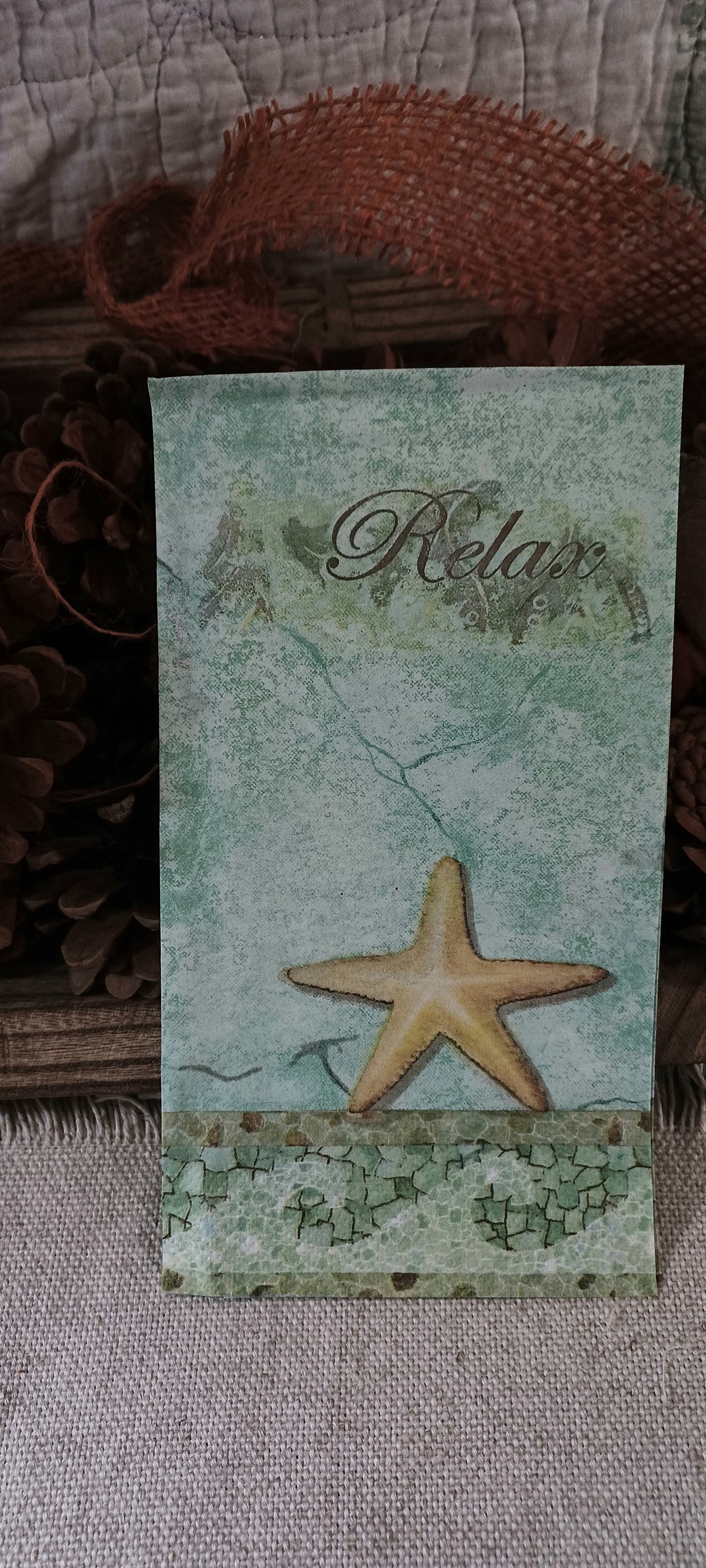 Styled photograph of an aqua vintage coastal guest napkin with golden starfish and Relax script displayed with rustic natural decor.