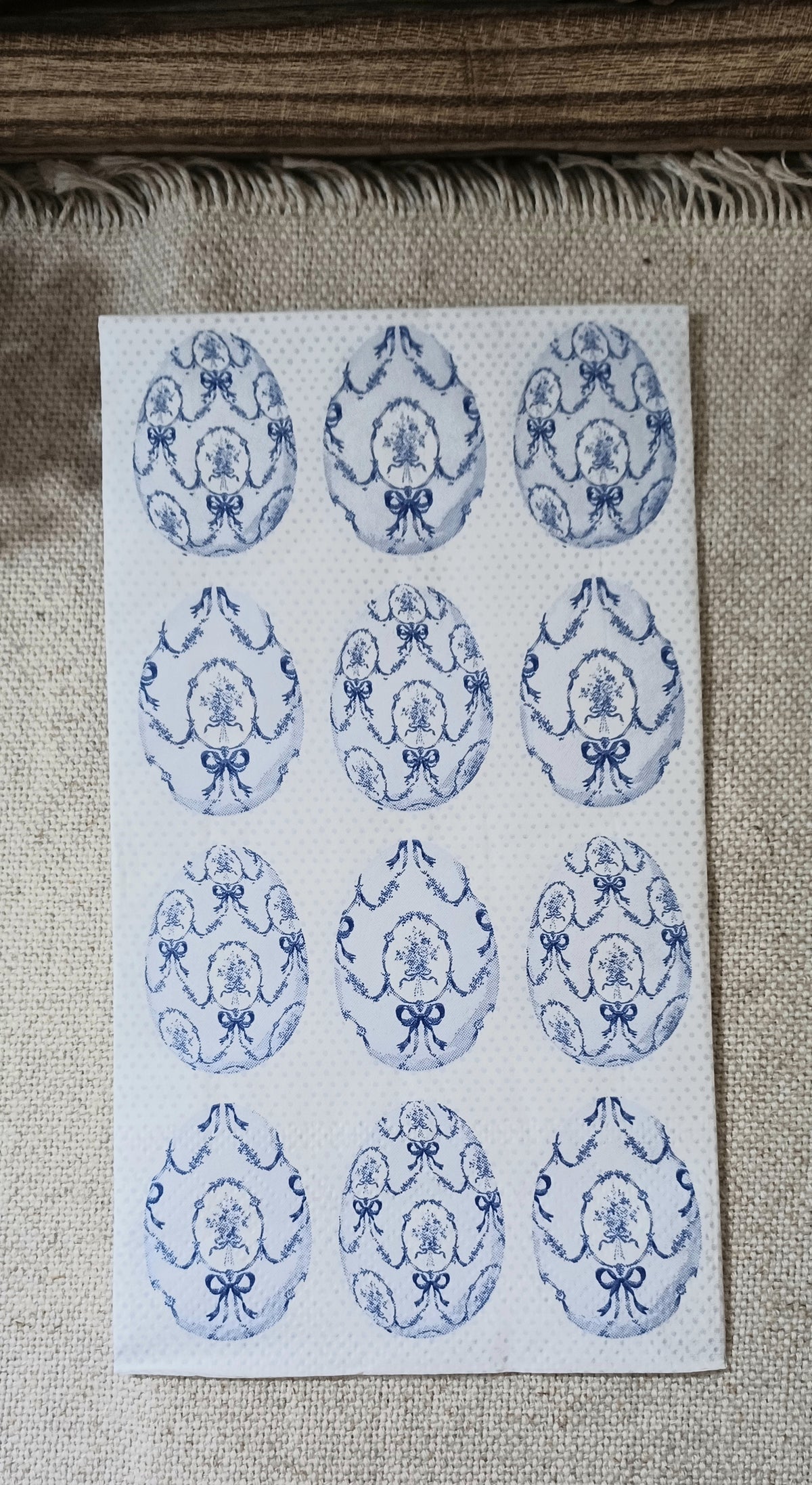 Blue and white paper guest napkin with repeating vintage-style oval floral medallions, bows, garlands, and a pale dotted background.
