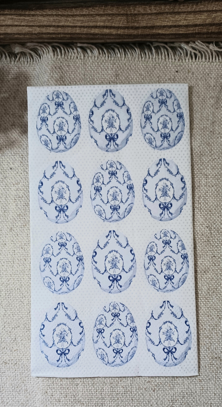 Blue and white paper guest napkin with repeating vintage-style oval floral medallions, bows, garlands, and a pale dotted background.