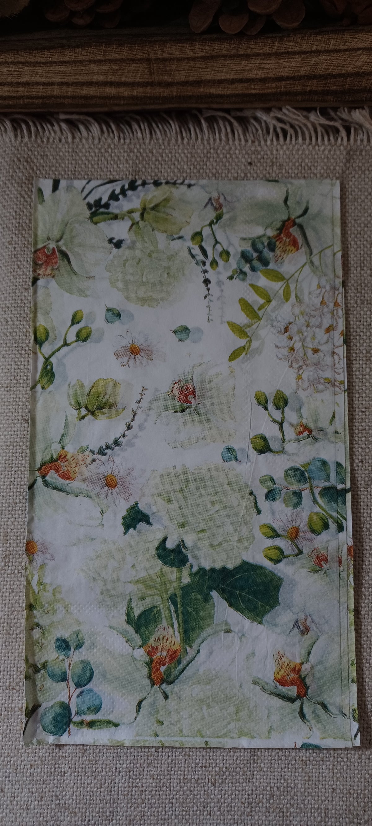 Watercolor floral guest napkin featuring delicate garden blossoms and muted green foliage displayed against a neutral fabric background.