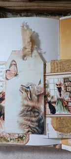 A close-up of a handmade scrapbook or collage page featuring a large printed kitten illustration, a butterfly, burlap textures, vintage-style paper elements, and a small illustrated figure in a room.