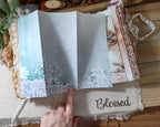 A paper accordion page or scrapbook panel with a white and teal floral design and lace border, held open by a hand over a rustic wooden surface with a 'Blessed' fabric underneath.
