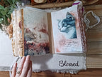 An open handmade journal with vintage-style cat illustrations and handwritten script, held by a hand over a frayed white cloth that says 'Blessed' on a wooden surface.