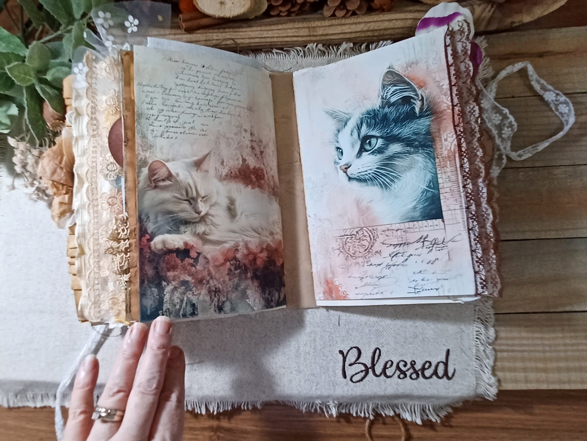 An open handmade journal with vintage-style cat illustrations and handwritten script, held by a hand over a frayed white cloth that says 'Blessed' on a wooden surface.