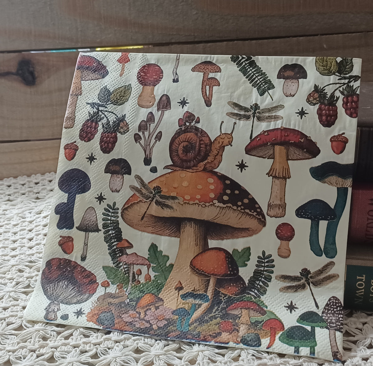 Woodland luncheon napkin featuring a large spotted mushroom with snail surrounded by mushrooms, dragonflies, and forest botanicals.