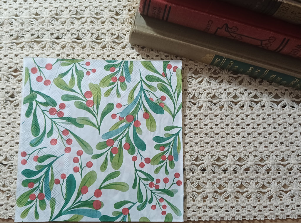 Botanical paper napkin with green watercolor foliage and red berries displayed upright beside vintage books.
