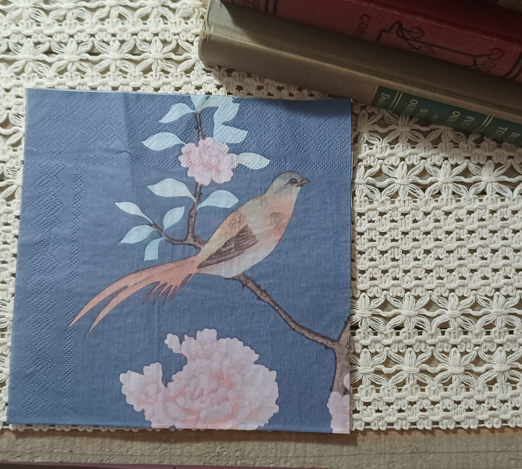 Blush bird and pink blossom paper napkin displayed upright beside vintage books on a crocheted textile.