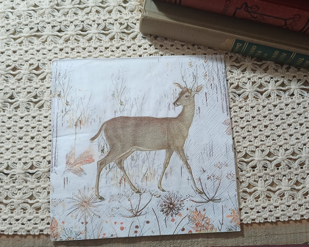 Vintage-style deer and winter meadow paper napkin displayed upright beside old books on a cream crocheted textile.