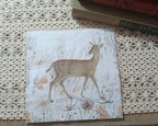 Vintage-style deer and winter meadow paper napkin displayed upright beside old books on a cream crocheted textile.