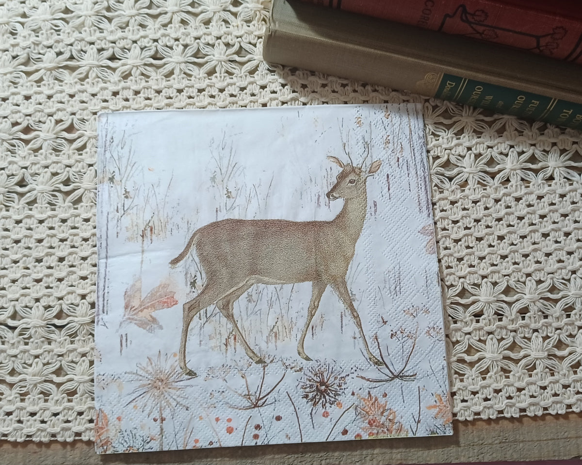 Vintage-style deer and winter meadow paper napkin displayed upright beside old books on a cream crocheted textile.