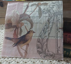 Vintage-style pink bird and old letters paper napkin displayed upright beside antique books on a cream crocheted textile.