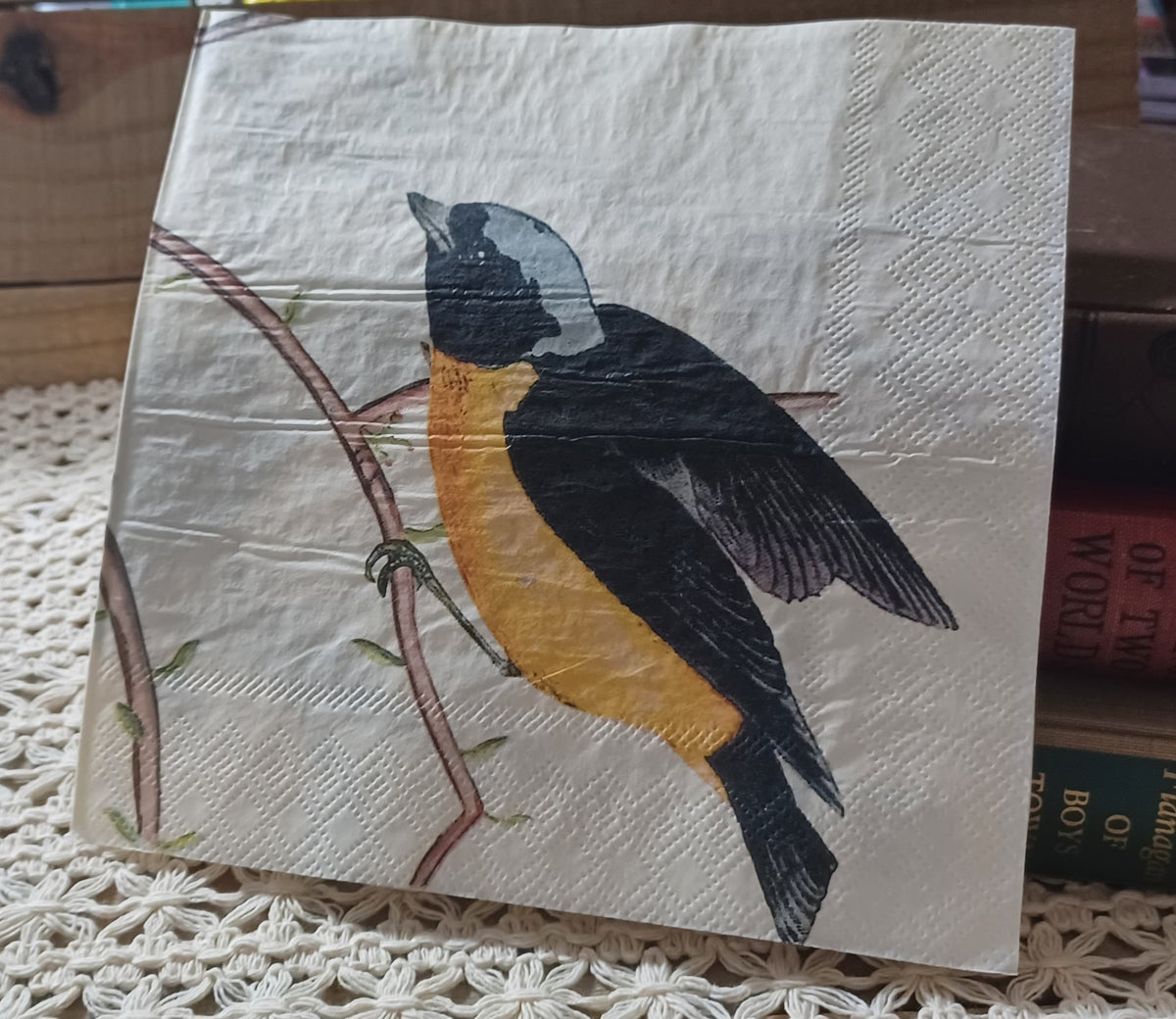 Golden-breasted songbird paper napkin displayed upright beside vintage books on a cream crocheted textile.