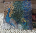 Vintage-style peacock paper napkin displayed upright beside old books on a cream crocheted textile.