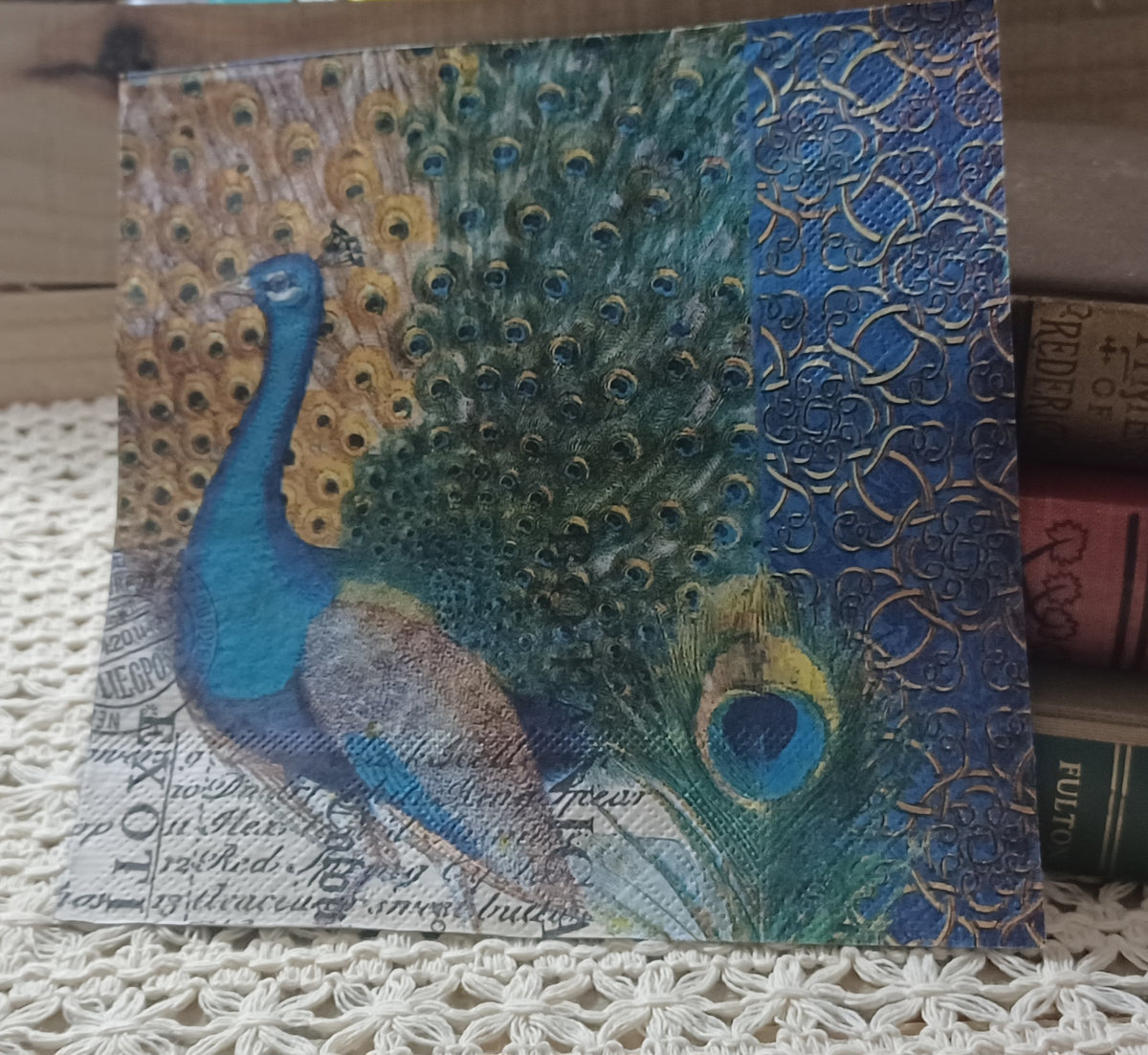 Vintage-style peacock paper napkin displayed upright beside old books on a cream crocheted textile.