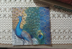 Jewel-tone paper luncheon napkin featuring a blue and green peacock with spread tail feathers, antique script and an ornate blue-and-gold border.