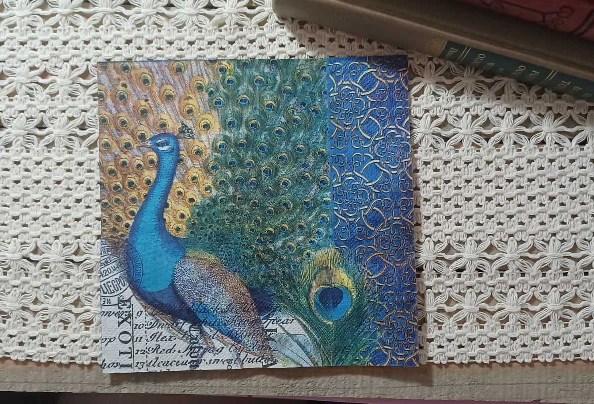 Jewel-tone paper luncheon napkin featuring a blue and green peacock with spread tail feathers, antique script and an ornate blue-and-gold border.