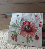 Decorative paper napkin featuring a large red ladybug on a pink flower with green leaves, smaller ladybugs, hearts, and floral details.