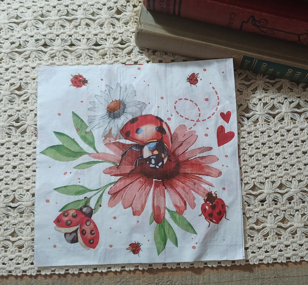 Full view of the floral side of a ladybug decorative paper napkin with a red ladybug, pink flower, white daisy, green leaves, hearts, and small garden insects.