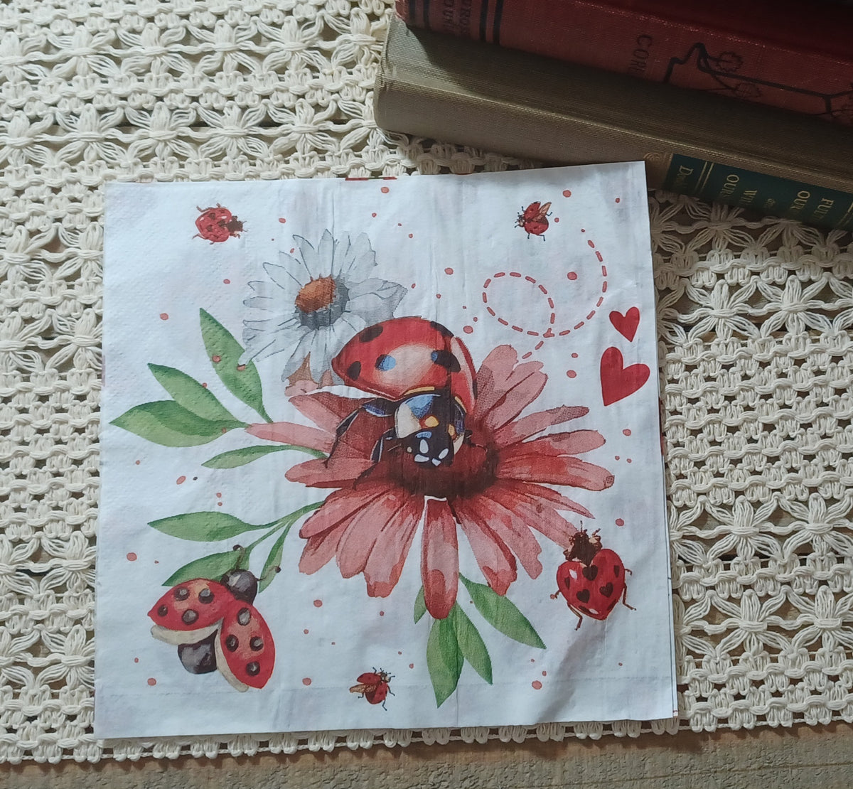 Full view of the floral side of a ladybug decorative paper napkin with a red ladybug, pink flower, white daisy, green leaves, hearts, and small garden insects.
