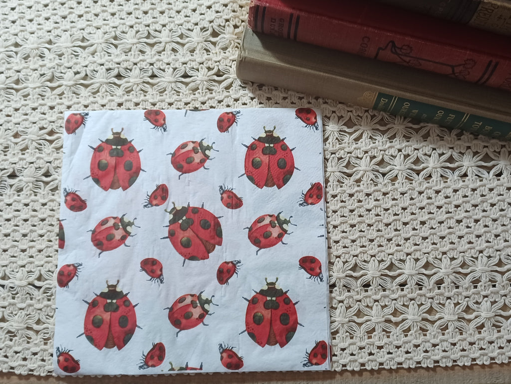 Overhead view of an individual decorative paper napkin with a repeating red and black ladybug pattern, photographed beside vintage books and crochet lace.