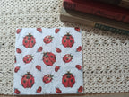 Overhead view of an individual decorative paper napkin with a repeating red and black ladybug pattern, photographed beside vintage books and crochet lace.
