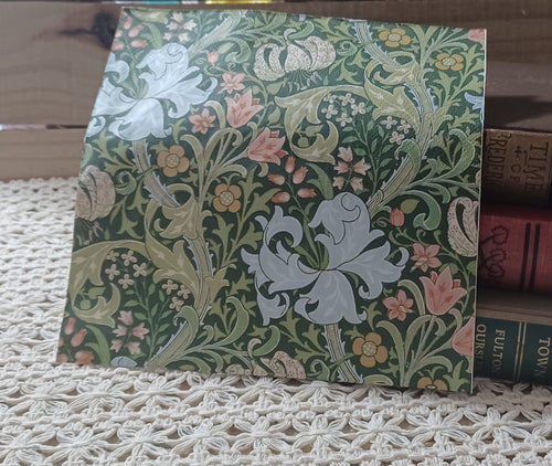 Overhead view of an individual botanical decoupage napkin with pale blue lilies, blush flowers, muted gold blossoms, and scrolling leaves on a deep green background.