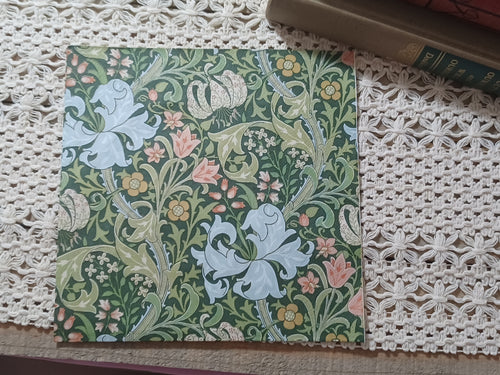 Decorative paper napkin with an ornate Arts and Crafts-inspired floral pattern featuring pale lilies, pink and gold flowers, and curling green foliage on a dark green background.