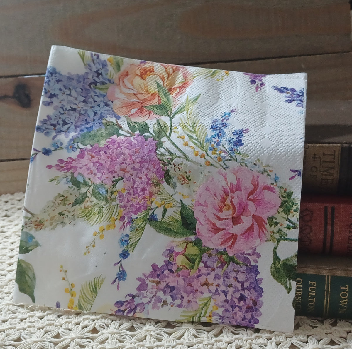 Overhead view of a single floral decoupage napkin with pink roses, lavender and purple flower clusters, small blue and yellow blossoms, and green leaves on a white background.