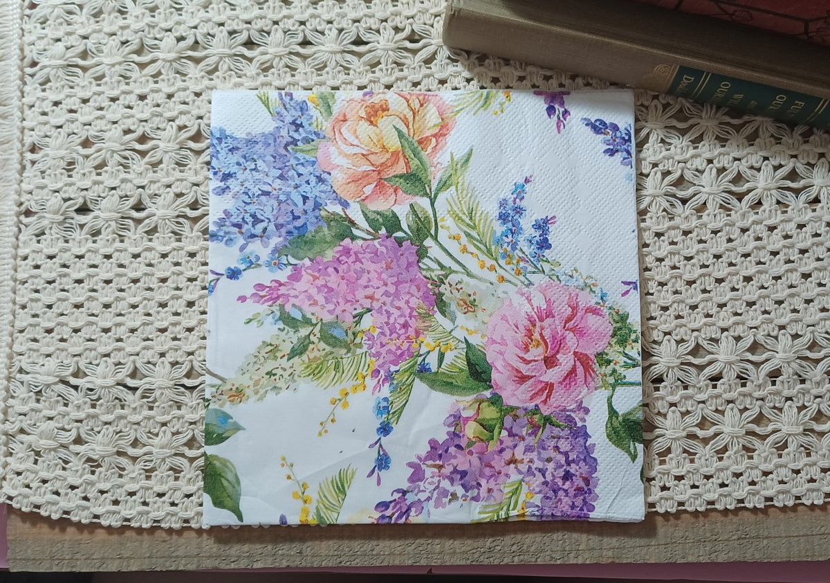 Decorative paper napkin featuring watercolor-style pink and peach roses, purple lilac flower clusters, blue blossoms, yellow sprigs and green foliage on a white background.
