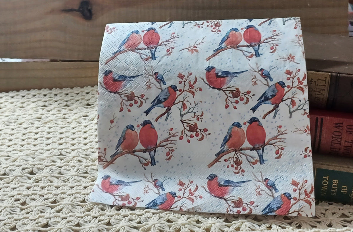 Overhead view of a decorative paper napkin patterned with red-breasted winter birds, berry-covered branches and small pale blue snow dots.