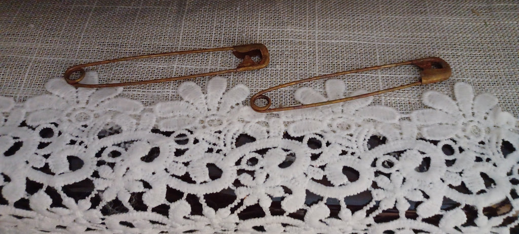 Two large 3½-inch metal safety pins showing varied reddish-brown genuine rust finishes on a lace-covered surface