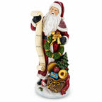 A Santa Claus figurine holding a scroll with names, decorated with a green wreath, gold bow, Christmas tree, ornaments, and gifts at the base, shown on a plain white background.