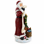 A Santa Claus figurine standing on a snowy base, holding a long scroll and next to a small decorated tree and gift bag, photographed on a plain white background.