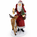 A Santa Claus figurine with a white beard, wearing a red coat with silver trim, holding a gold bell and a small green Christmas tree, standing next to a brown reindeer on a white base against a white background.