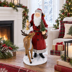 A festive Christmas figurine of Santa Claus standing with a reindeer on a snowy base, placed on a wooden table in a decorated living room with a fireplace, Christmas tree, and wrapped gifts.